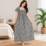 A woman stands barefoot in a bedroom wearing a long gray nightgown with a white floral print. The dress has short flutter sleeves, a smocked bodice, and a gathered skirt.