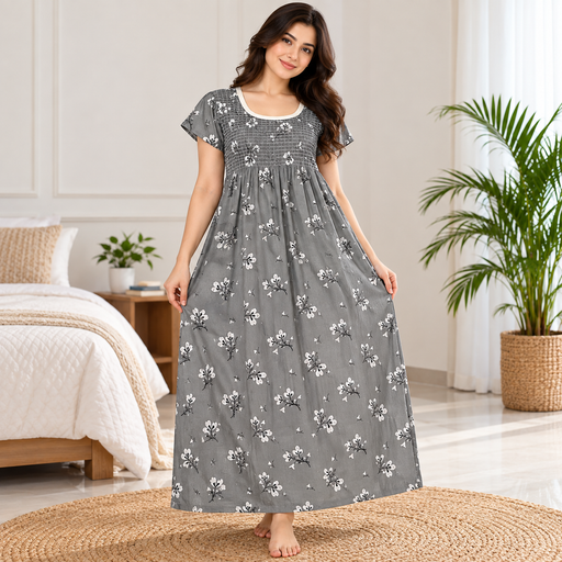 A woman stands barefoot in a bedroom wearing a long gray nightgown with a white floral print. The dress has short flutter sleeves, a smocked bodice, and a gathered skirt.