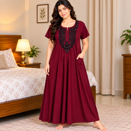 A woman stands barefoot in a bedroom wearing a long maroon nightgown with black embroidered detailing around the neckline and side pockets. The room features a wooden bed, bedside lamp, and neutral-toned curtains.